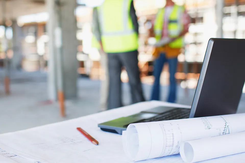 Architect drawings and laptop on construction site with background crew